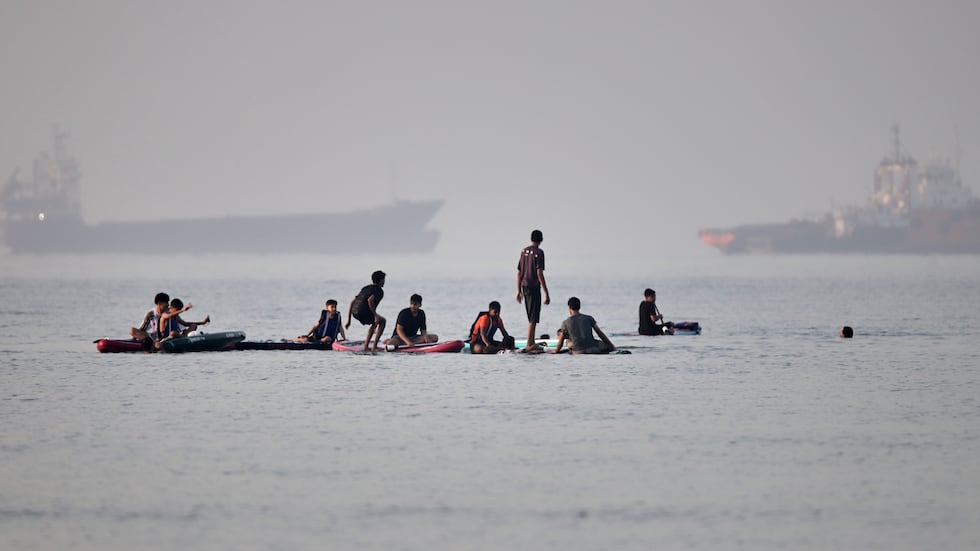 People gather on paddleboards in shallow water as cargo and service vessels are anchored in...