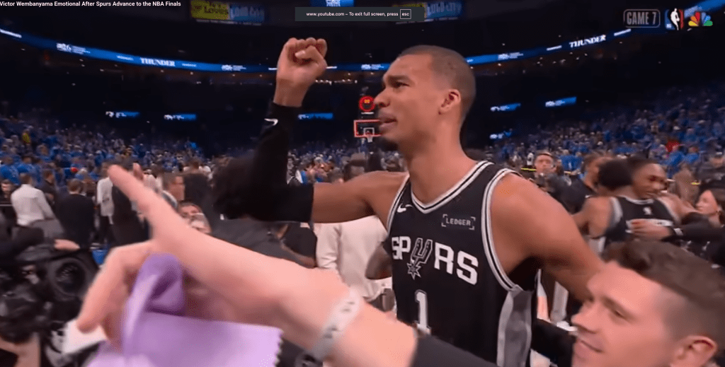 A man in a Spurs jersey raises his fist in a stadium full of people.