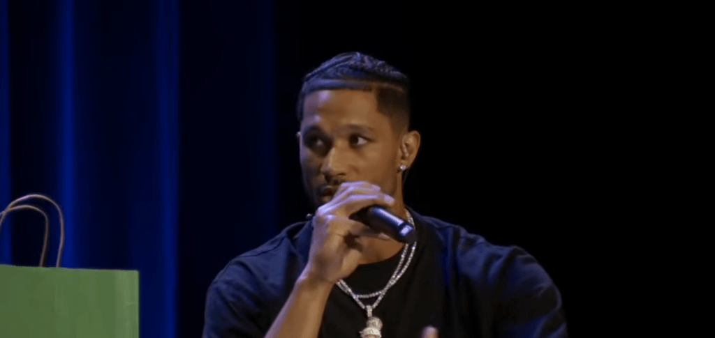 A man with braided hair, a dark shirt, and a shiny necklace is speaking into a microphone.