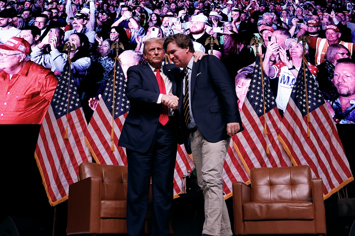 Donald Trump and Tucker Carlson stand on a stage shaking hands. President Trump on the left wears a dark suit, a white collared shirt, and a bright red tie, and has his left arm resting on the other man's shoulder. Tucker Carlson on the right wears a dark blazer, a striped tie, and khaki pants, and is leaning forward slightly with a smile. Behind them are several United States flags on poles, two brown leather armchairs, and a large digital screen displaying a crowded audience of people watching.