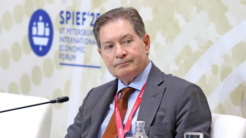 Man with brown and greying hair, wearing a grey suit with blue short and brown tie. A red lanyard is around his neck.
