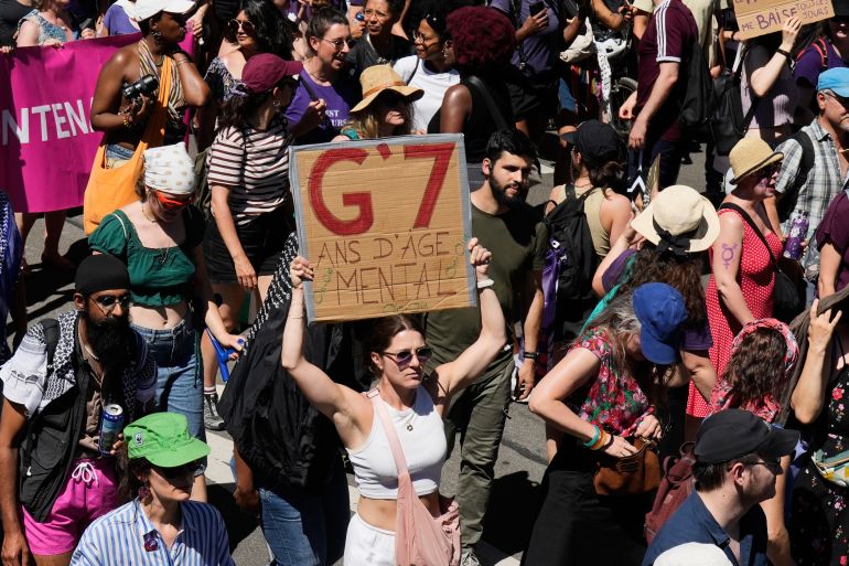 A woman holds a sign which reads "G7-mental age years" as she participates in a "No G7" demonstration in Geneva, Switzerland, Sunday, June 14, 2026, ahead of the G7 summit scheduled to take place June 15-17. (AP Photo/Baz Ratner)