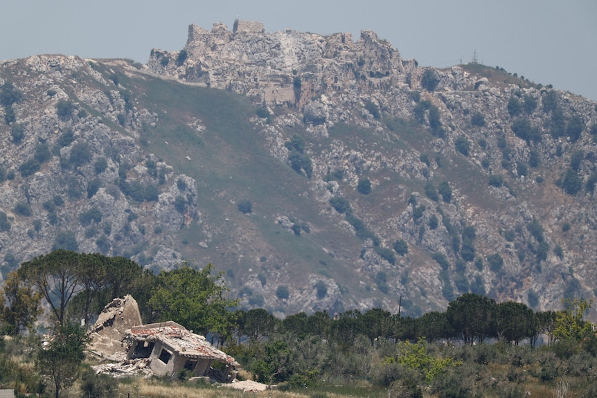 Crumbled ruins of a medieval castle atop a giant mountain dotted by greenery, behind a lower foreground of trees.