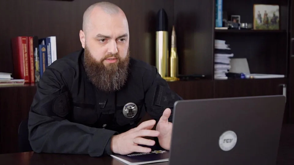 Vitaliy Sova, a man with a shaved head and a long brown beard, speaking on a video call in his laptop while he is filmed from a different angle in the office, which has black bookshelves behind him.