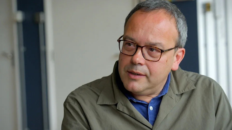 Nick Lowles, a man with greying hair and dark-rimmed glasses, wearing a grey-green jacket over a blue shirt