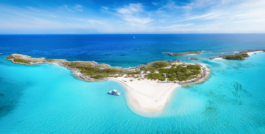Panoramic aerial view of the Exumas Cays in The Bahamas, showing turquoise waters, white sandbars, and small islands with lush vegetation, with a white boat anchored near a sandy beach.