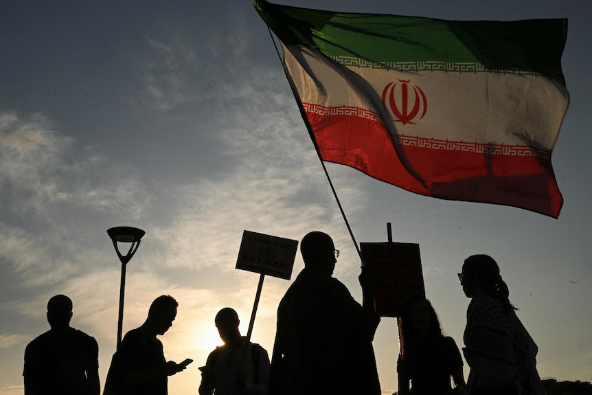 The official Iranian flag is waving in the wind above demonstrators who are in the shadows.