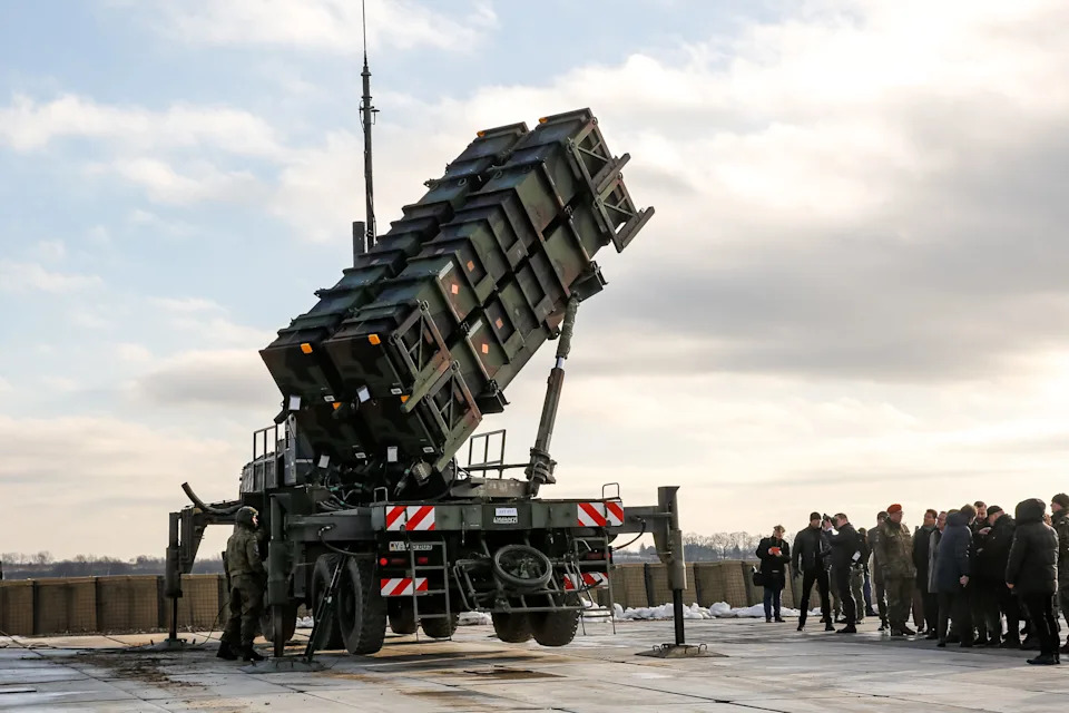 A large camouflage-coloured defense system on the back of a truck points into the sky with figures standing around it