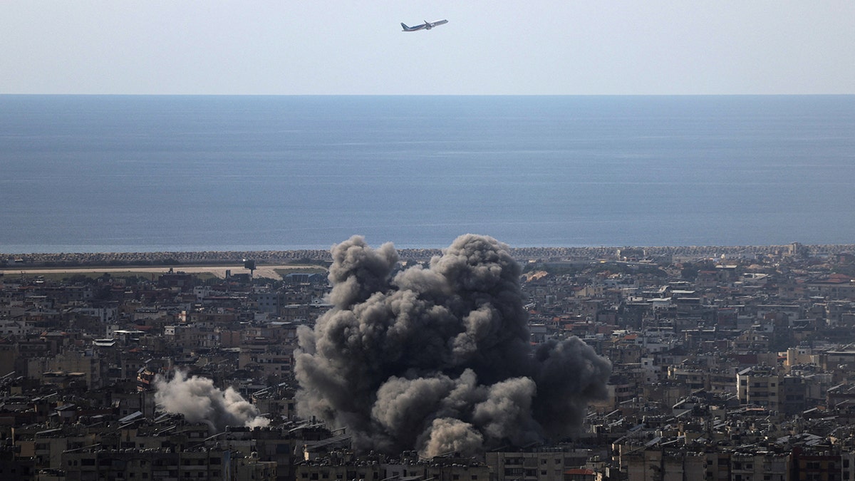 An airplane flies over Beirut's southern suburbs with smoke rising from the ground
