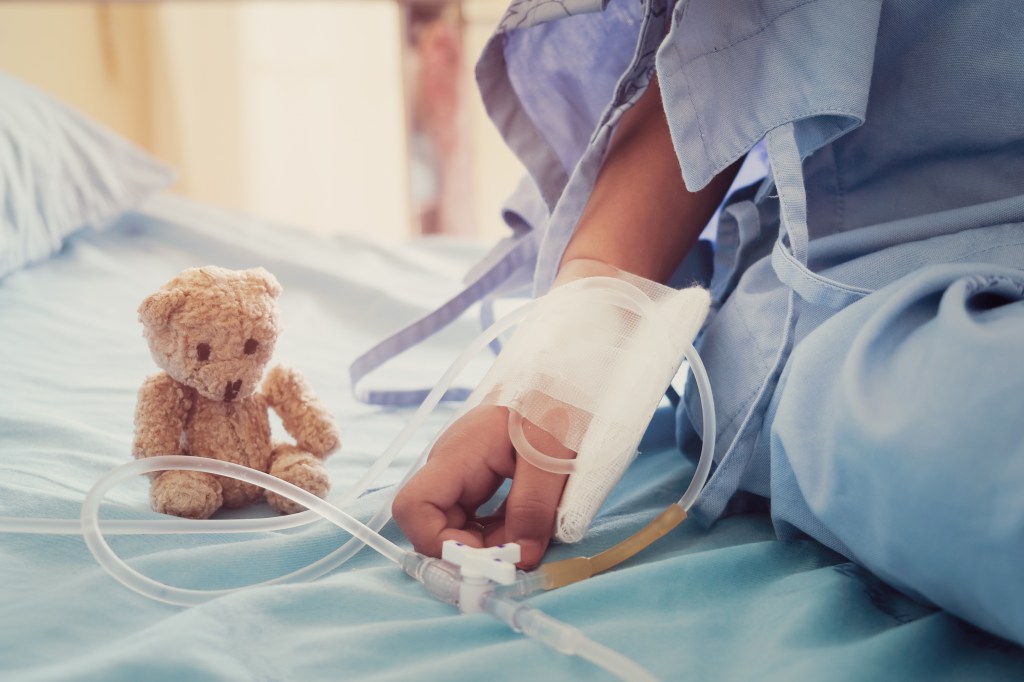 A child in a hospital bed with an IV in their hand, next to a teddy bear.