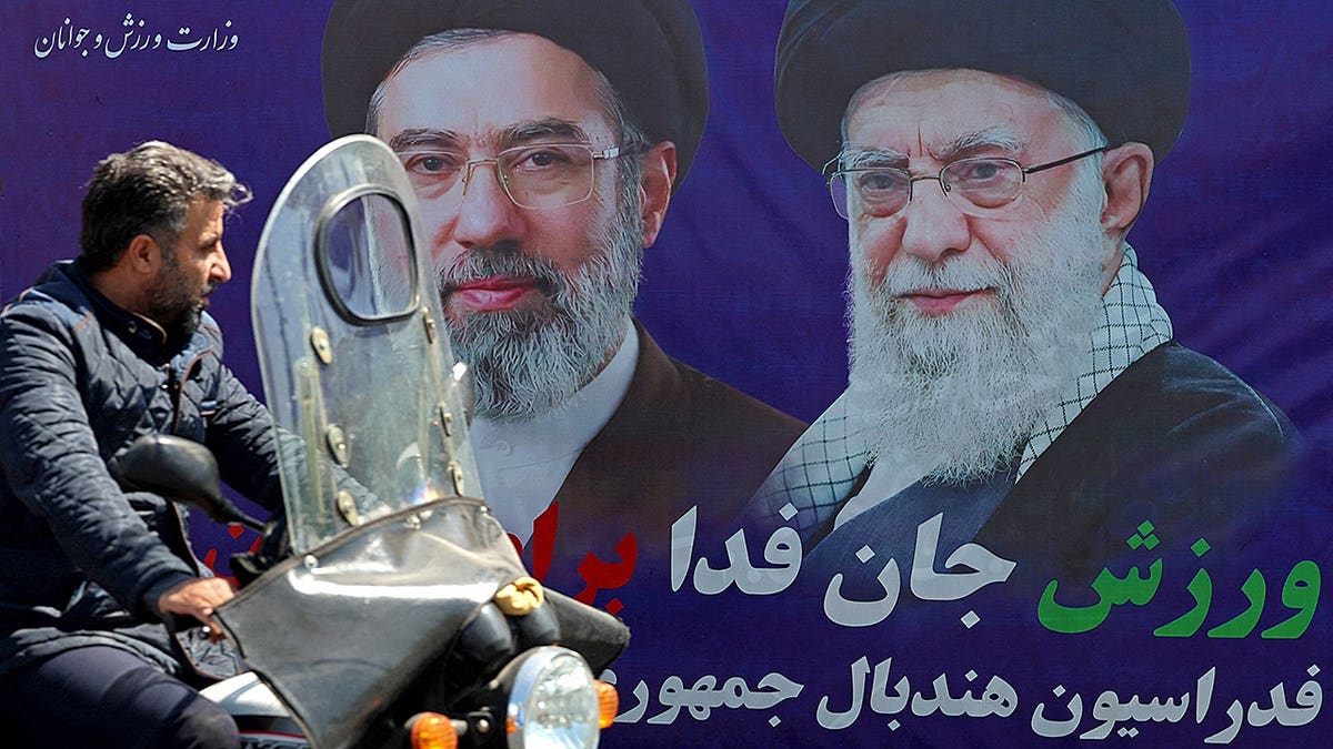 A motorist riding past a banner with images of Ayatollah Ali Khamenei and Mojtaba Khamenei in Tehran