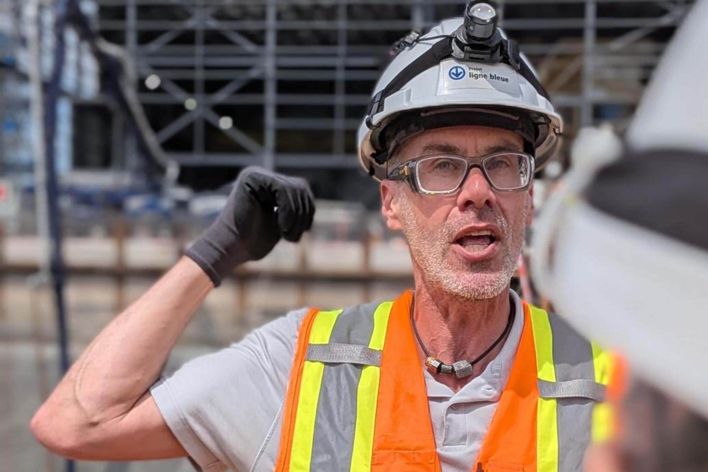 David Chartier gestures on a construction site while wearing a hard hat and reflective vest.