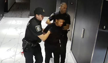 Two police officers walk a man in handcuffs to an elevator.