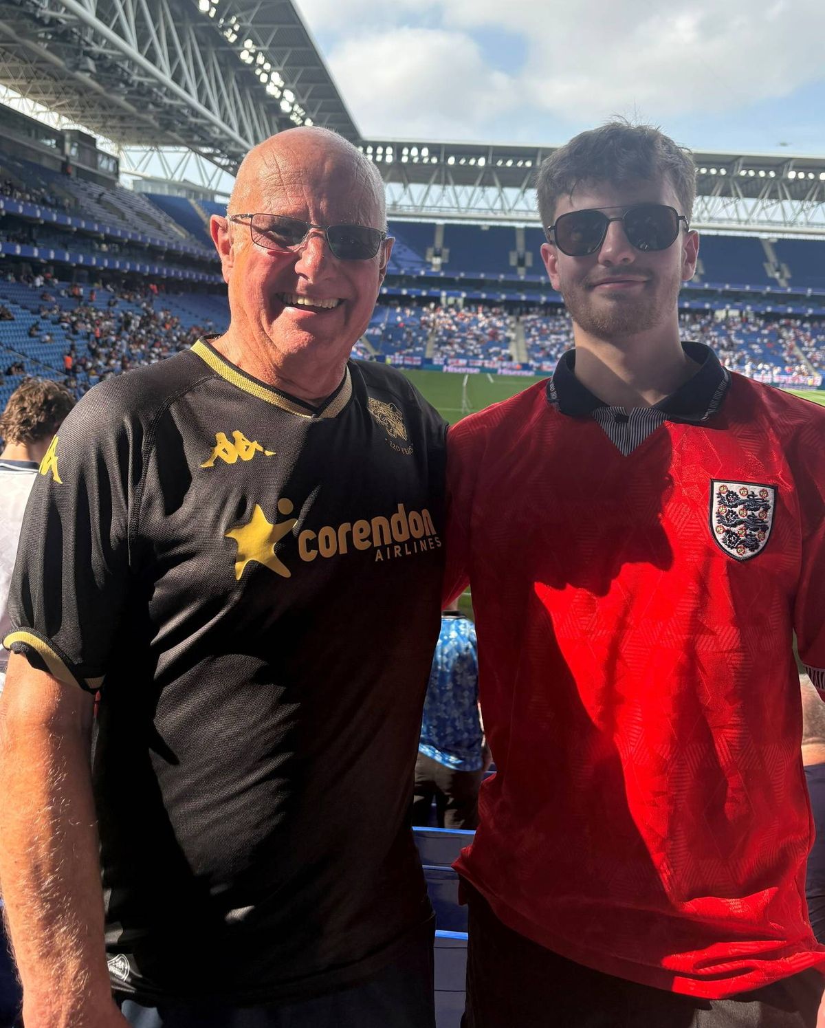 Two individuals, one wearing a black and yellow jersey with a logo and the other in a red jersey, are standing side by side in front of a stadium with a crowd of spectators.