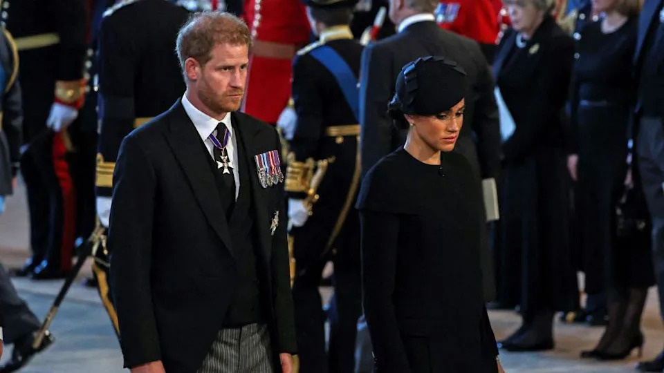 Mid-shot of Prince Harry and Meghan Markle at the Queen's funeral in 2022. Prince Harry is wearing a black morning suit with a white shirt and black tie with military medals on his left side. Meghan is wearing all black.
