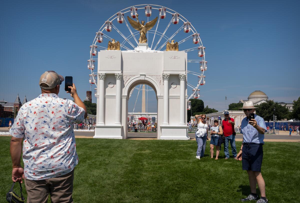 A man takes a photo of a model of an arch near a Ferris wheel.