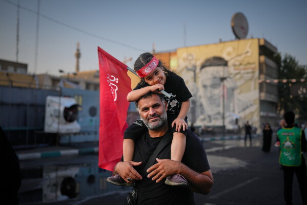 A man carries a child holding a red Shiite religious flag outside the Imam Khomeini Mosalla Grand Mosque during the funeral ceremonies for slain Iranian Supreme Leader Ayatollah Ali Khamenei and members of his family in Tehran, Iran, Saturday, July 4, 2026. (AP Photo/Altaf Qadri)