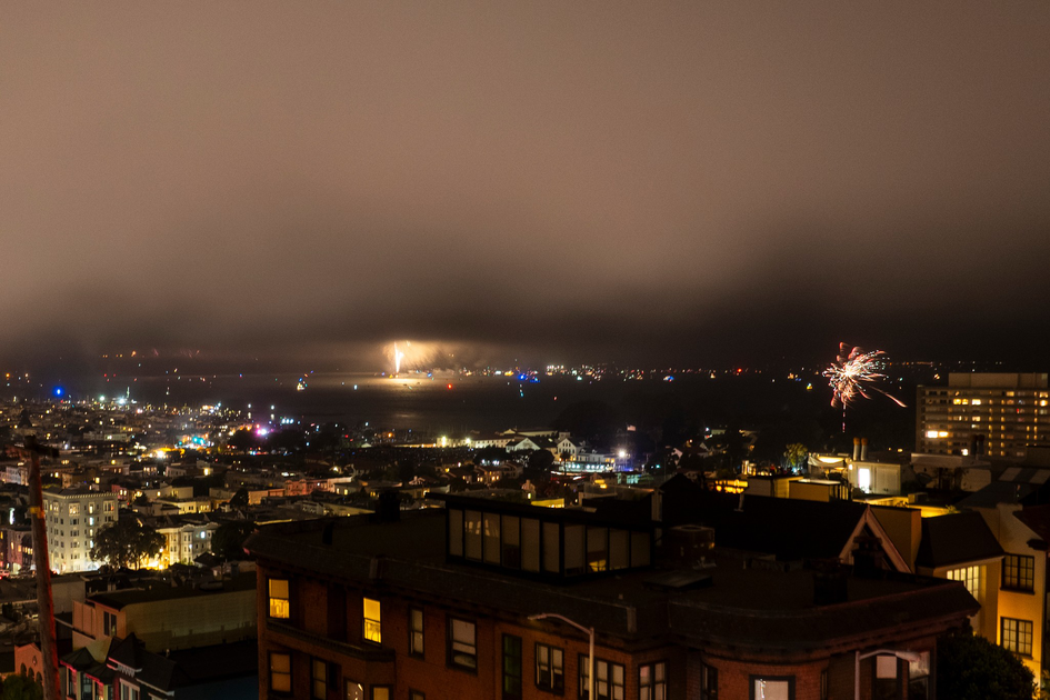 A cityscape at night includes fireworks bursting over a body of water, with buildings illuminated and a cloudy sky overhead.