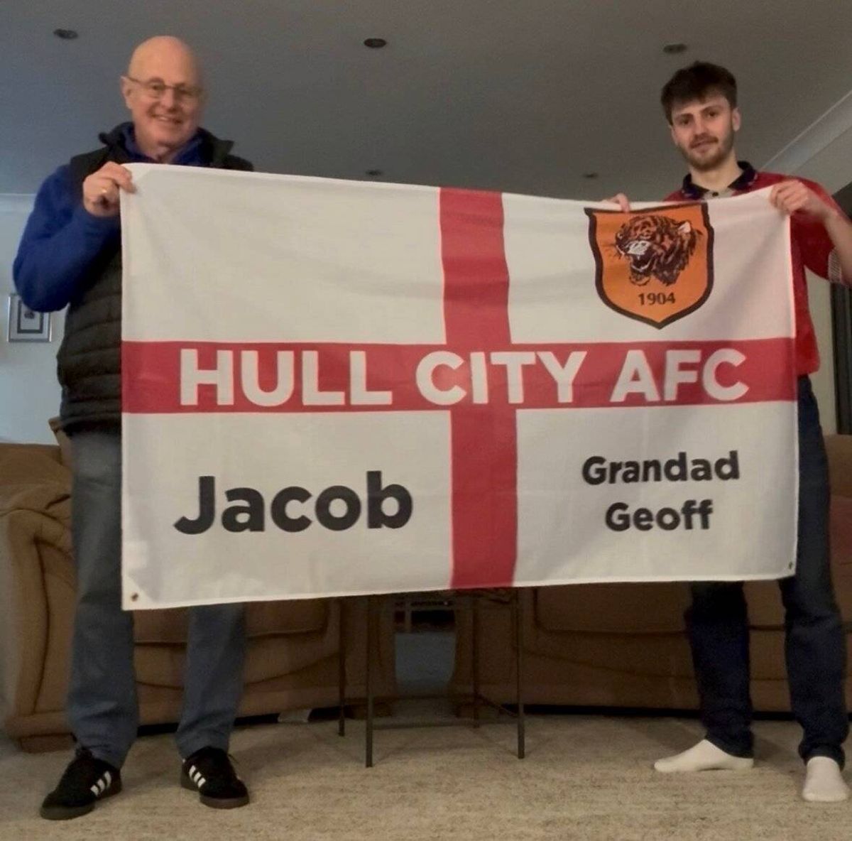 Two individuals are standing and holding a banner that reads "HULL CITY AFC" in large white letters on a red and white background. The banner features the names "Jacob" and "Grandad Geoff" in black text. The man on the left is dressed in a blue jacket, and the man on the right is wearing a red shirt.