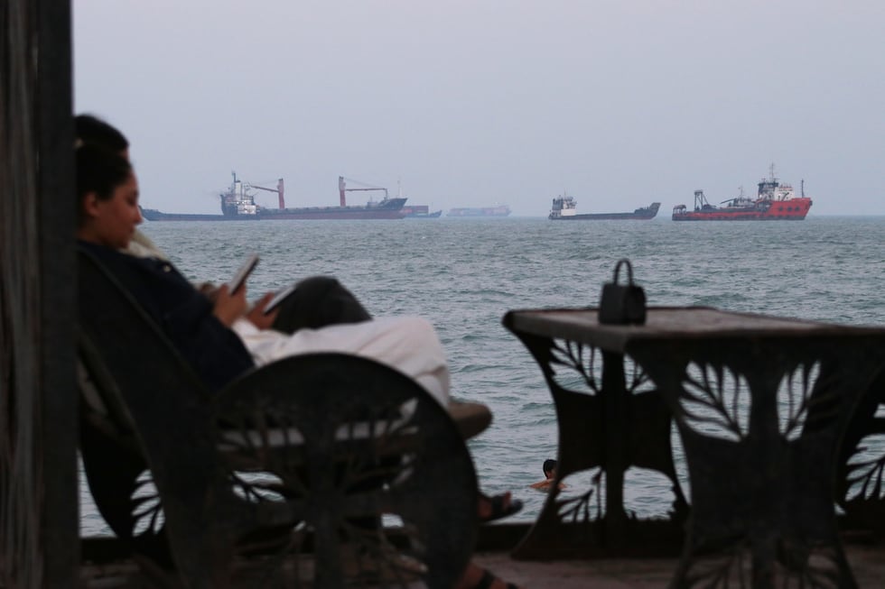 Residents check their cellphones as they sit at a cafe overlooking commercial vessels anchored...