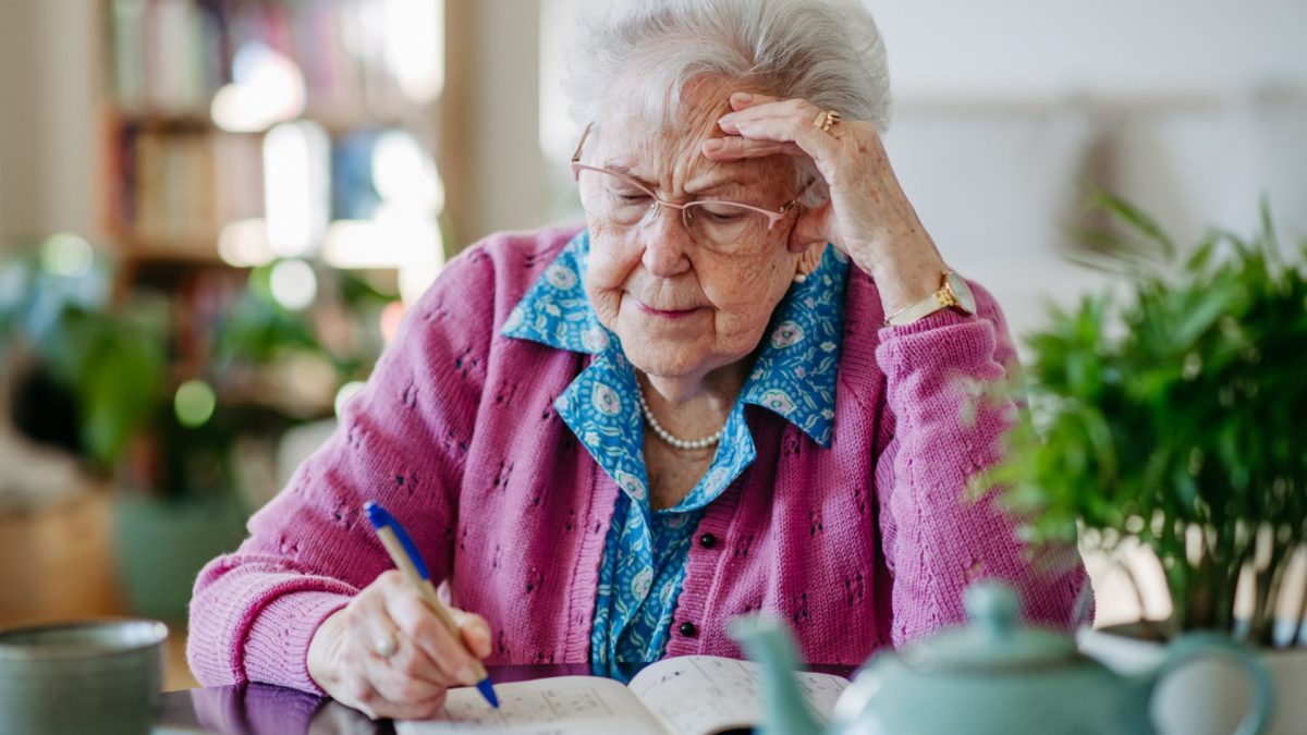 Portrait of elderly woman solving sudoku puzzles at home, sitting in living room. Sudoku as popular game for aging people, logical thinking, problem solving.