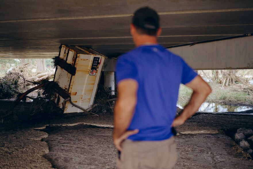 A man stands under a bridge looking a large metal box that is stuck underneath.