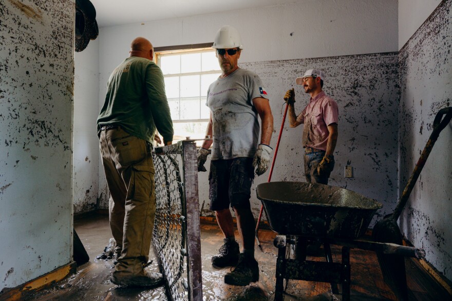 Three men stand inside a room ,with lines on the walls from flooding, while holding cleaning and moving supplies including a wheelbarrow.