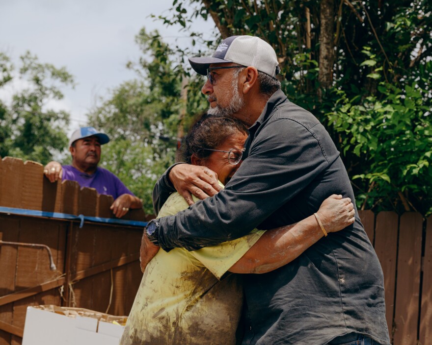 A man hugs a woman who is crying while another man looks over a fence in the background.