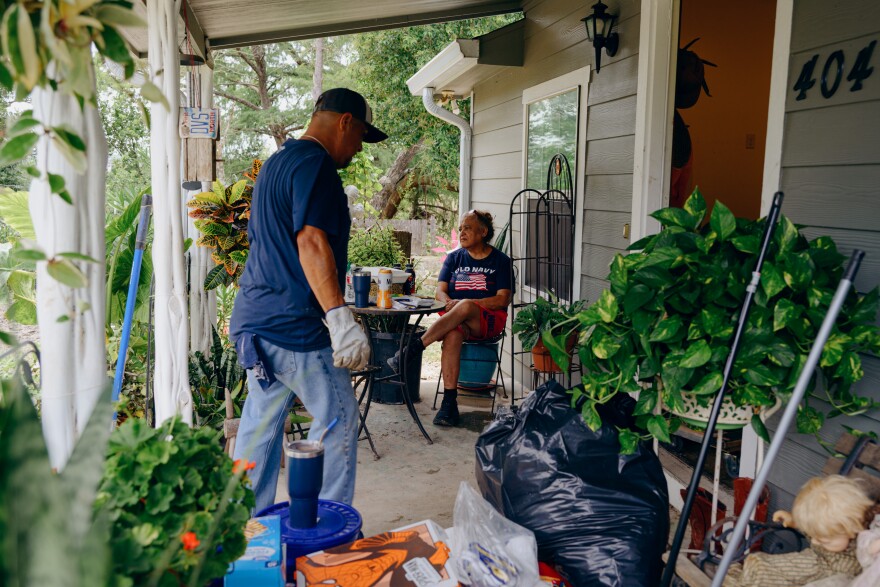A woman sits on her porch which has bags on it while a man walks by heading inside.