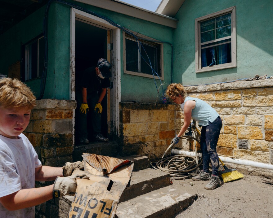 A woman uses a pressure washer to clean off a set of steps outside a home as a boy carries away trash on the left and another boy stands in the doorway.