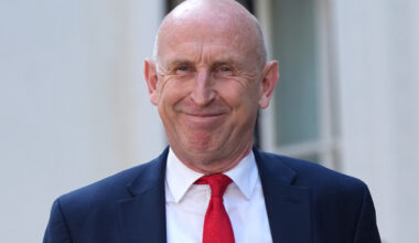 Chancellor John Healey smiling, wearing a navy suit, white shirt, and red tie.