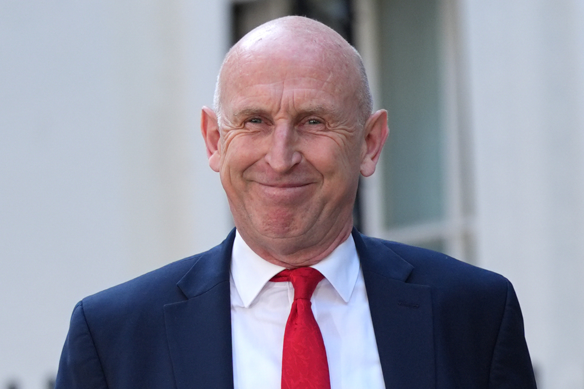 Chancellor John Healey smiling, wearing a navy suit, white shirt, and red tie.