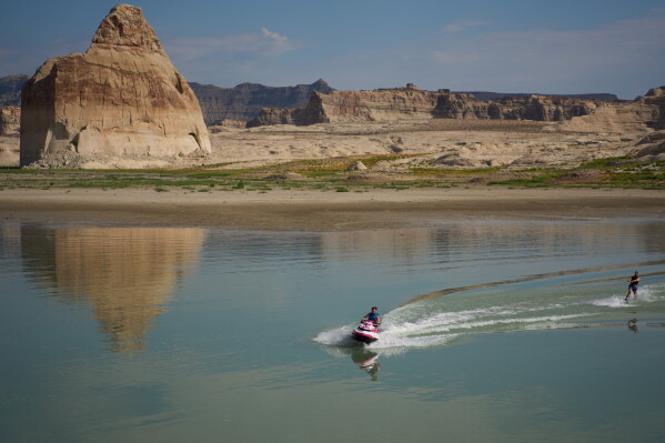 Vitalii Volna drives a jet ski on Lake Powell at the Glen Canyon National Recreation Area, Wednesday, July 15, 2026, near Big Water, Utah. A bathtub ring of light minerals on Lone Rock behind shows the high water mark of the lake. (AP Photo/John Locher)
