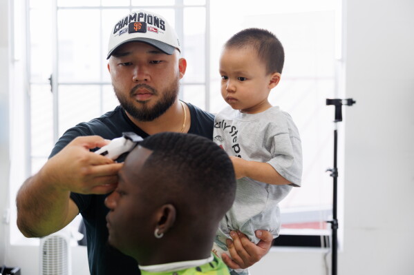 Jack Chang cuts Isaac Obamehinti's hair while holding his 2-year-old son Cairo Miu at his studio on Monday, July 13, 2026, in San Francisco. (AP Photo/Juliana Yamada)