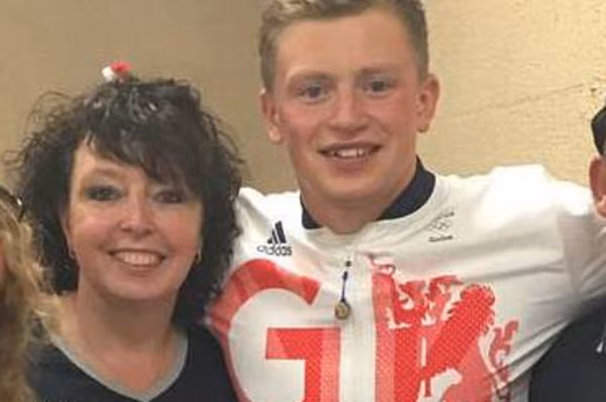 In an indoor setting, a young man dressed in a white and red football jersey with the letters "GK" prominently displayed on the front is posing for a photograph. Standing beside him, a woman with curly hair smiles warmly, while another individual, partially visible, appears to be engaging in the photo session as well. The background is neutral, focusing attention on the individuals.