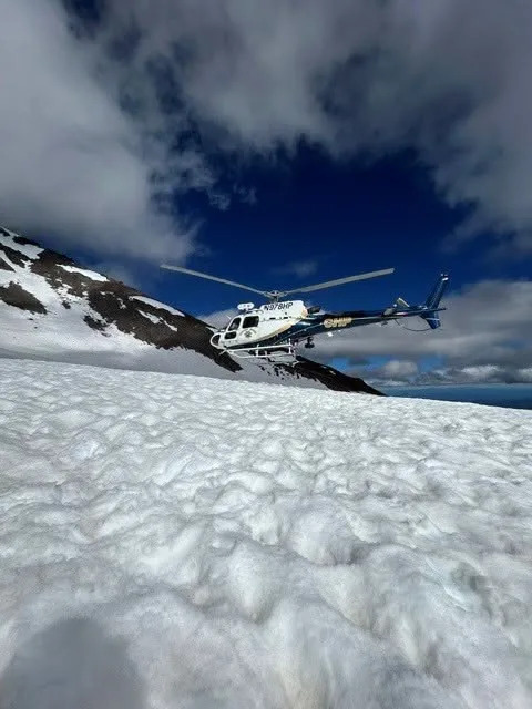 A rescue helicopter hovers over an icy, snow-covered mountainside under a cloudy blue sky. Facebook/U.S. Forest Service – Shasta-Trinity National Forest