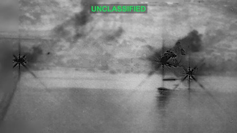 A column of smoke rises into the sky over a dark landscape in grainy aerial video footage