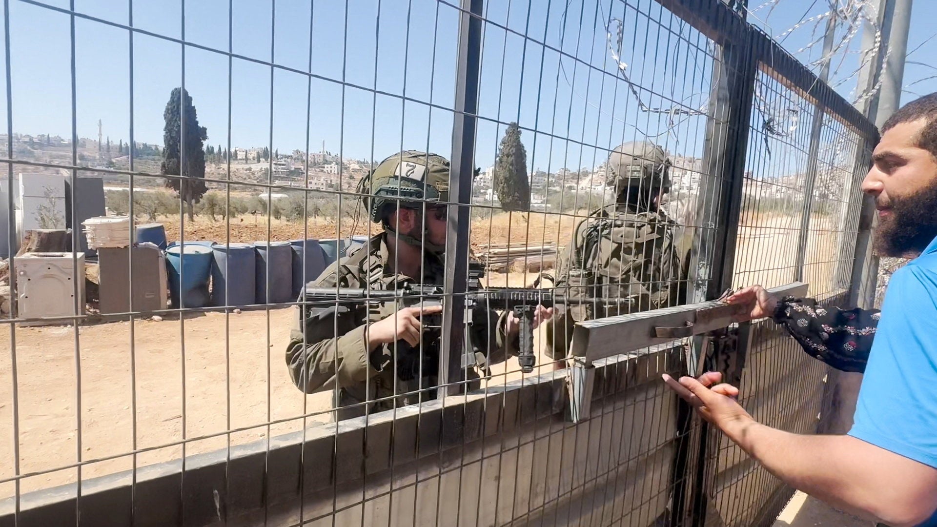 People wearing a military uniform stand behind a fence as a Palestinian man speaks to them from inside his home, during an Israeli settlers' attack on a Palestinian property in the village of Al-Mughayyir, near Ramallah, in the Israeli-occupied West Bank, July 11, 2026
