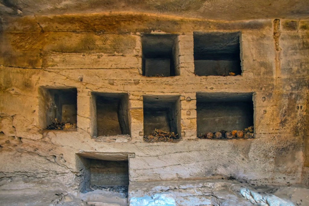 Interior of a limestone tomb with several niches containing skeletal remains and skulls.
