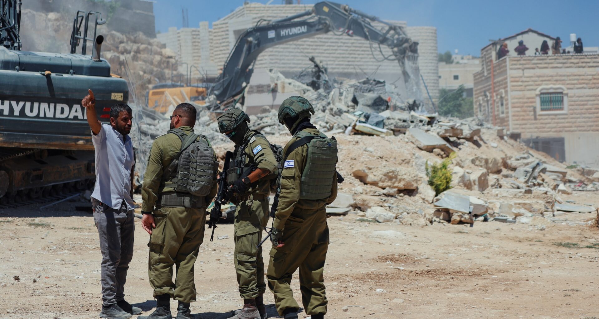 Israeli forces stand near an excavator as a Palestinian home is demolished in Al-Deirat, south of Hebron, occupied West Bank on July 14, 2026. Israeli forces demolish the home of Osama Masaaf on the grounds that it lacked an Israeli-issued building permit, displacing a Palestinian family of eight.