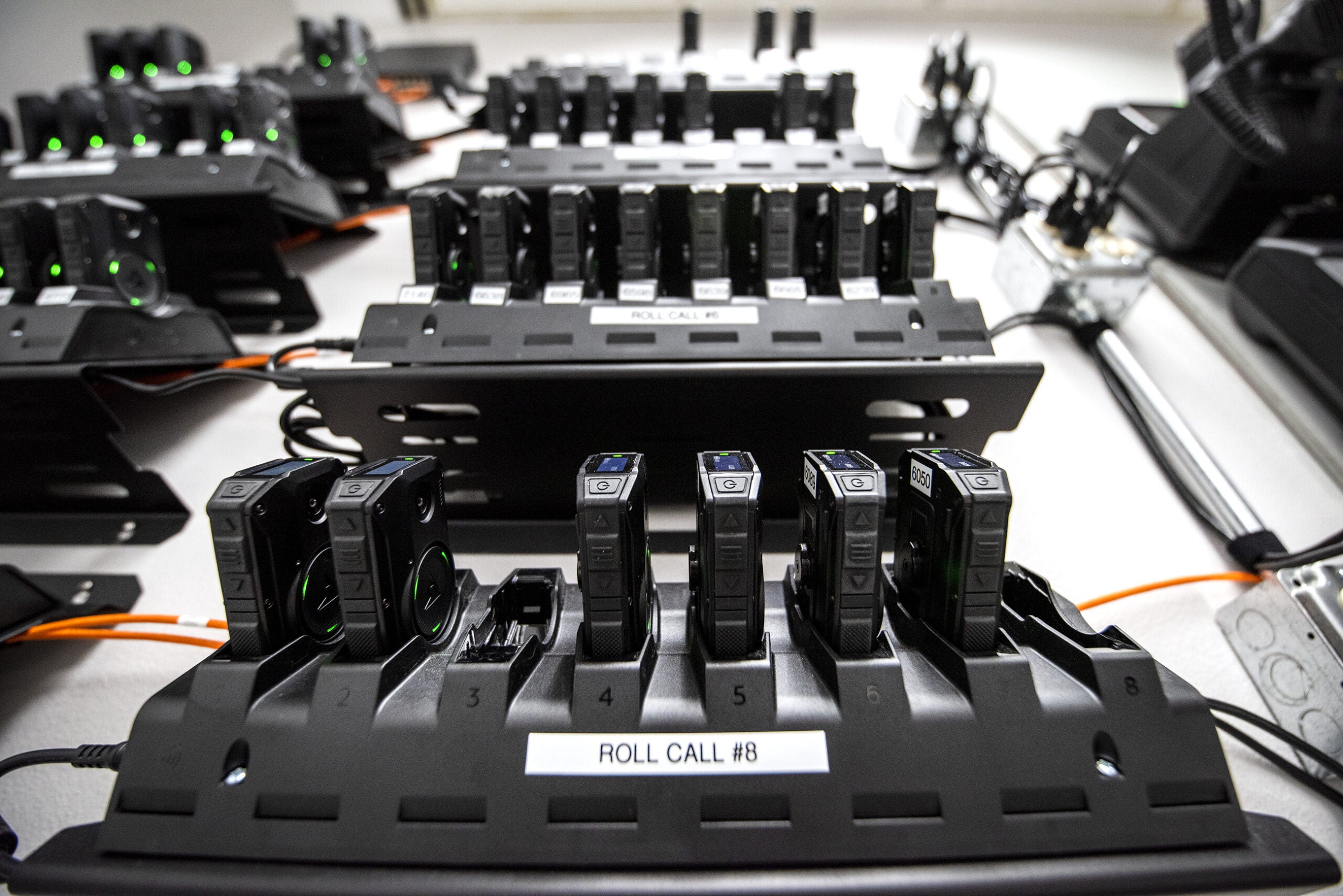 Rows of body cameras are docked onto black charging stations that are mounted to a white wall.