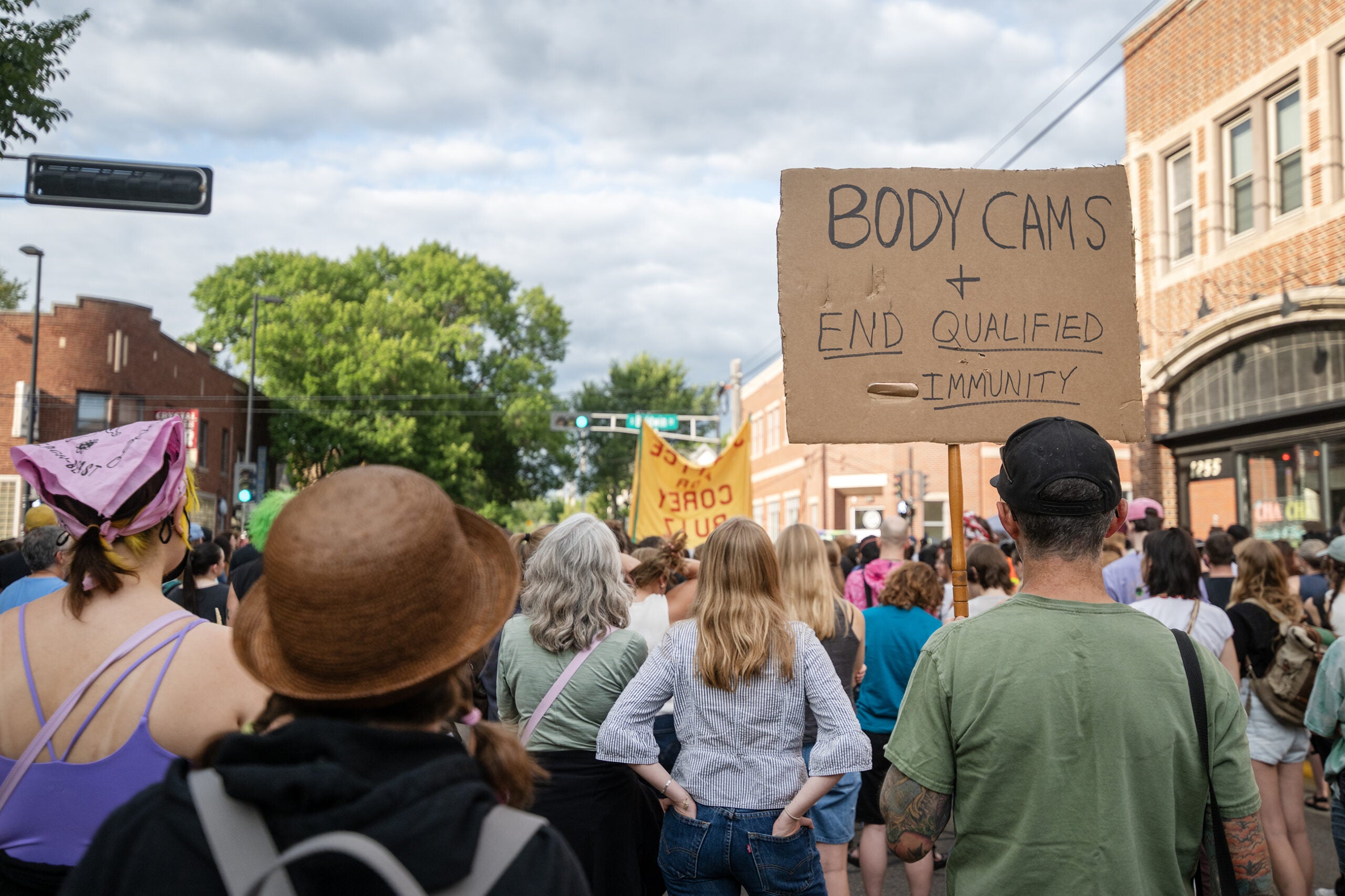 A group of people at a protest; one person holds a sign reading BODY CAMS + END QUALIFIED IMMUNITY. Buildings and trees line the street.