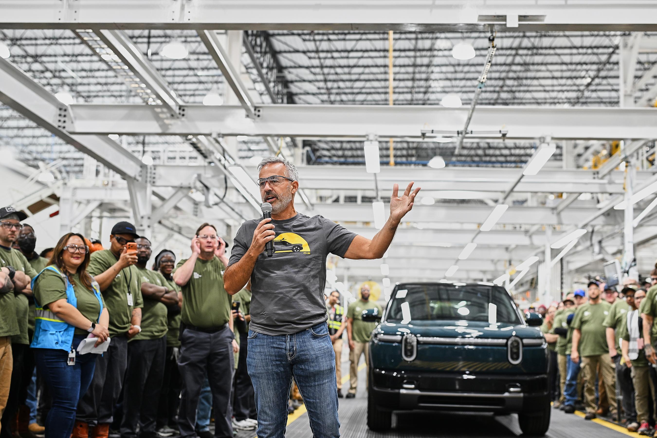 A man in a grey t-shirt and jeans holds a microphone and stands in front of a blue Rivian r2 inside of a huge, bright factory space and speaks to a crowd of people who are mostly wearing green t-shirts.