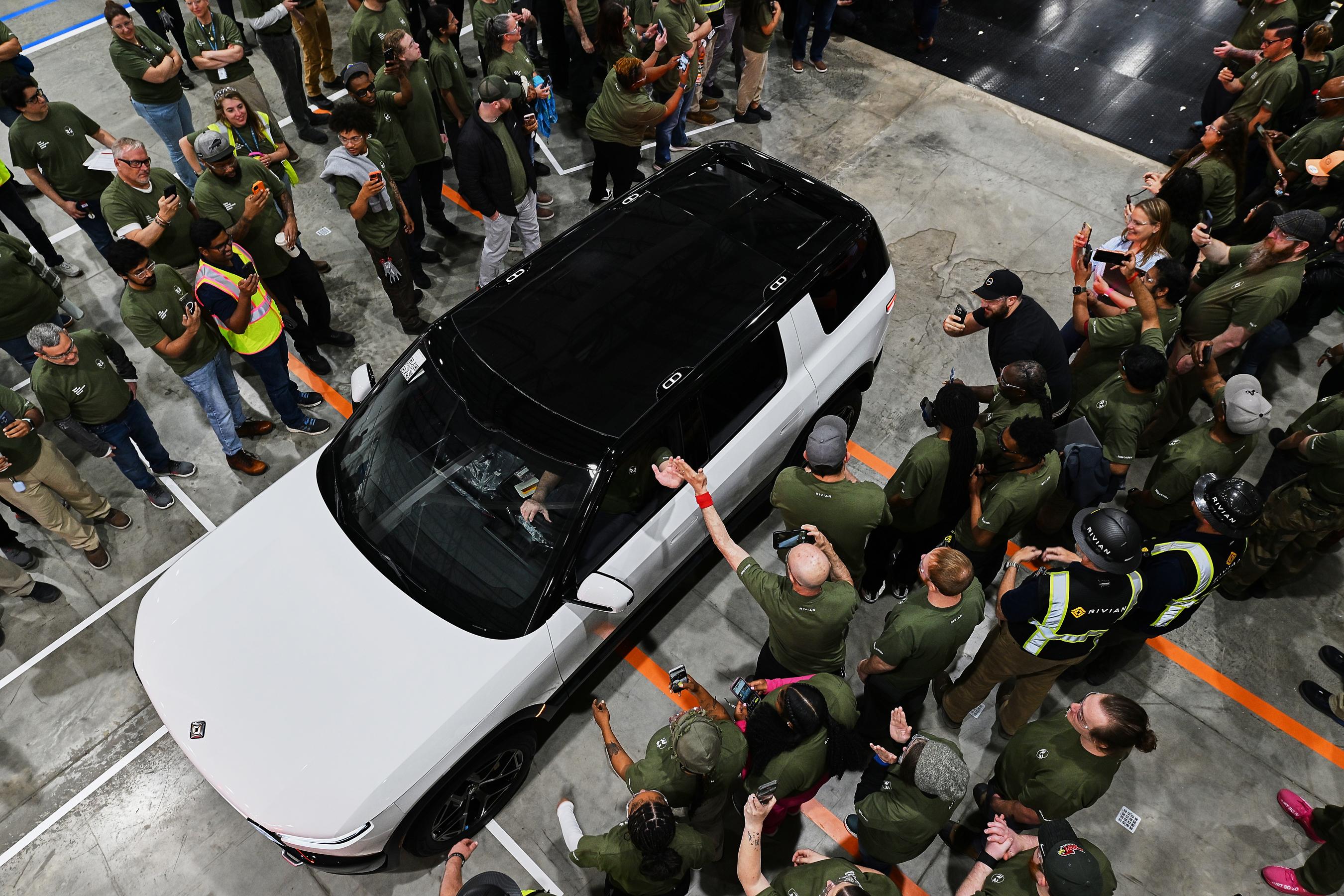 People lined up on either side of a white Rivian driving down a narrow path in a factory. The driver gives a high five to a person taking pictures outside of the vehicle.