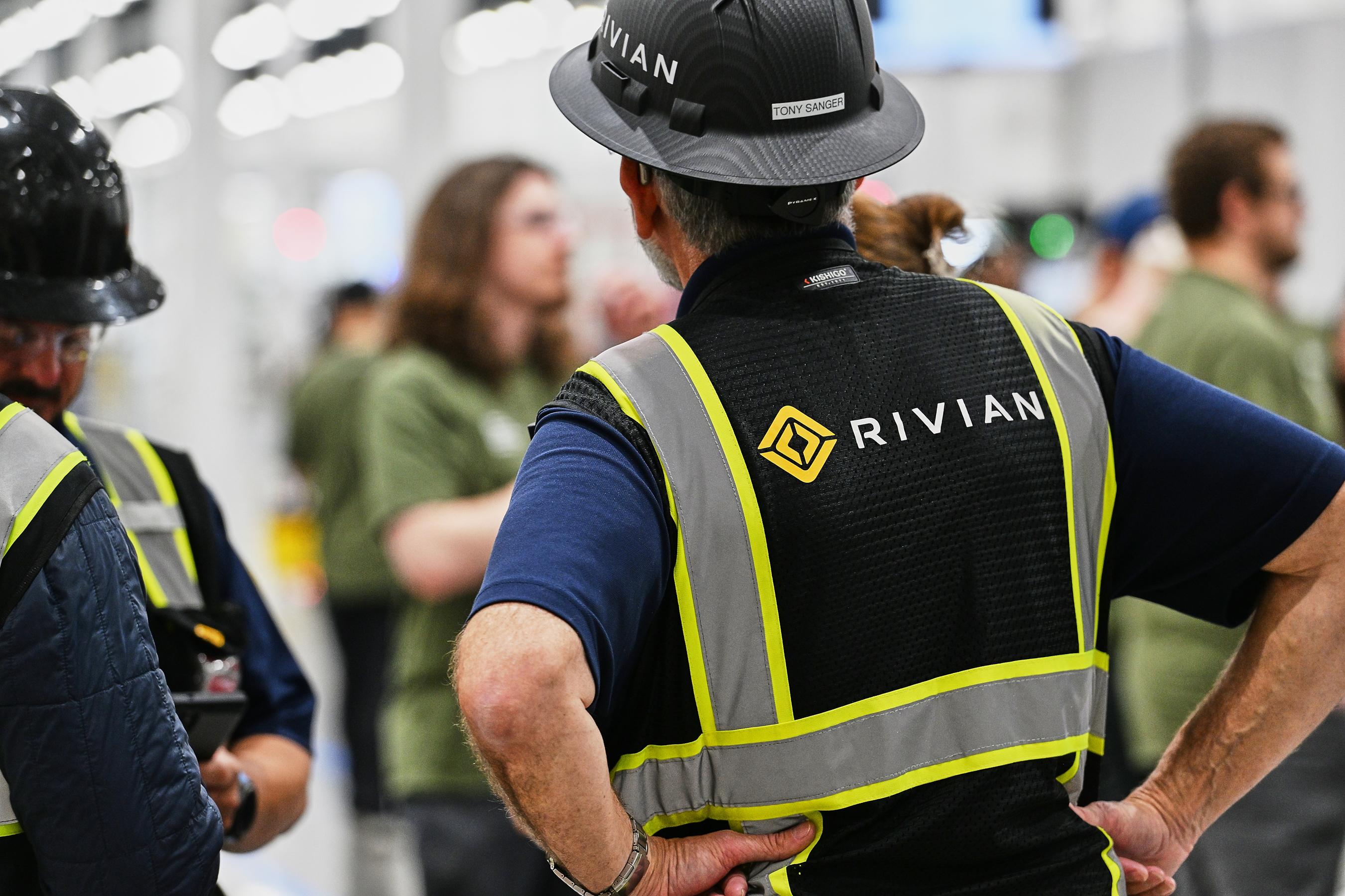 A person wears a black and yellow reflective vest with Rivian written on the back.