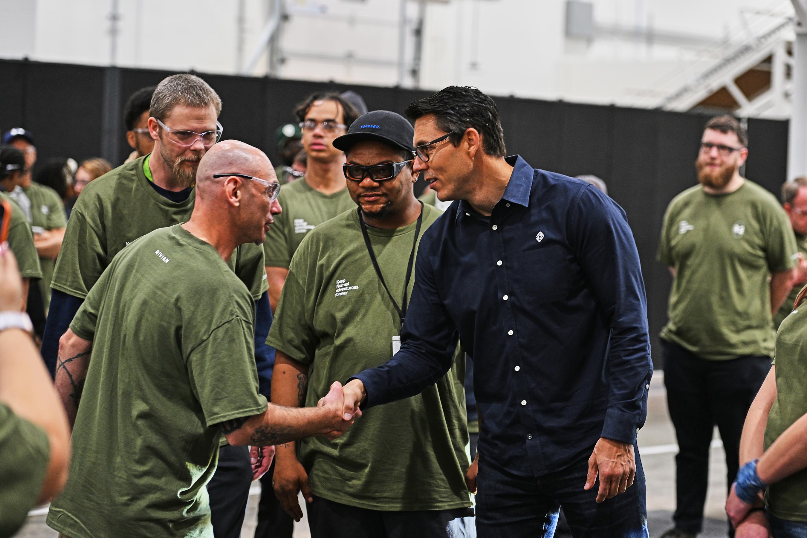 CEO RJ Scaringe in a blue long sleeved shirt and glasses is shaking the hand of a man in safety goggles and a green t-shirt. A group of people also in green t-shirts are around them.
