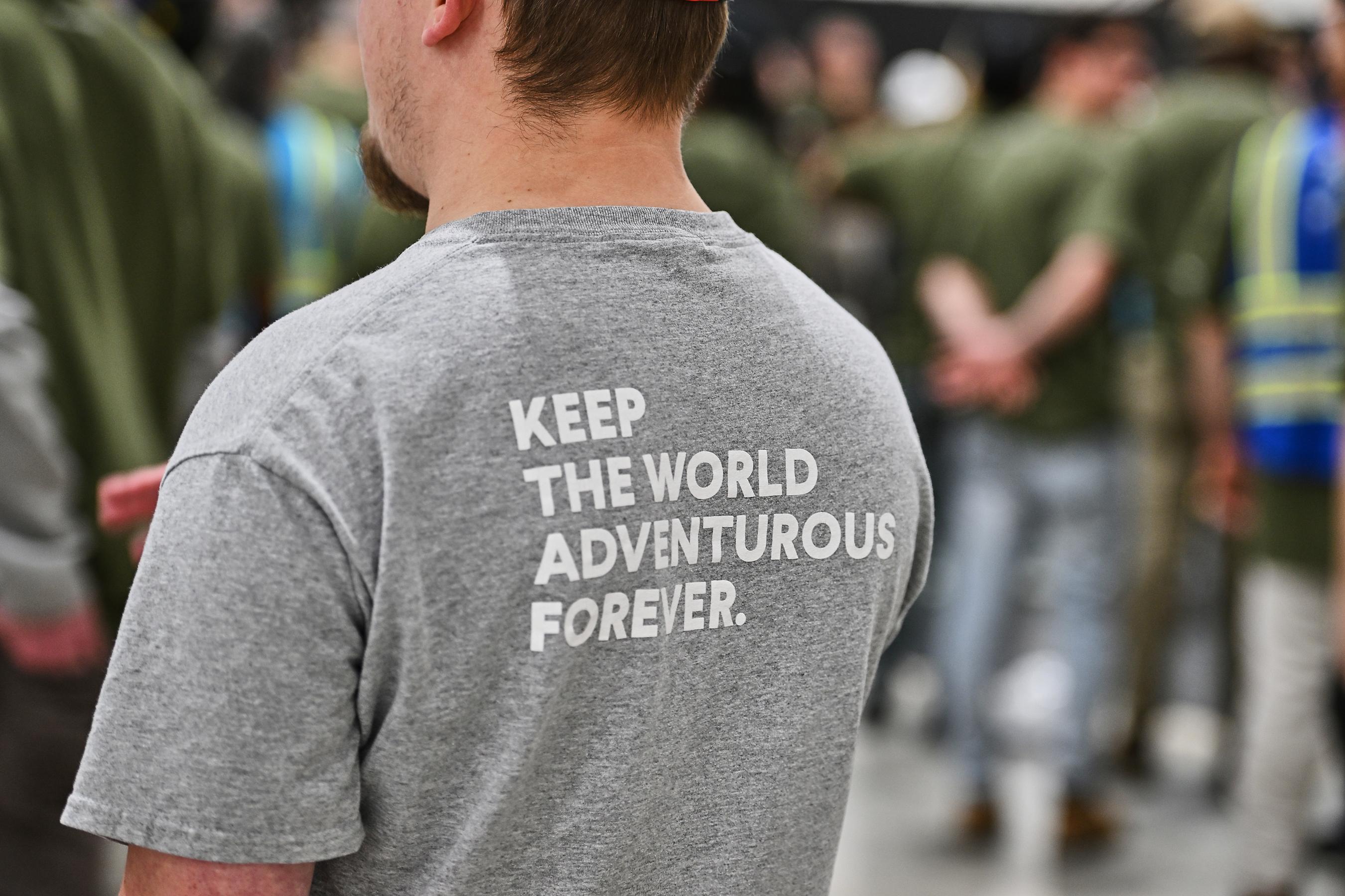 A person with short hair wears a gray shirt that reads “keep the world adventurous forever” on the back.
