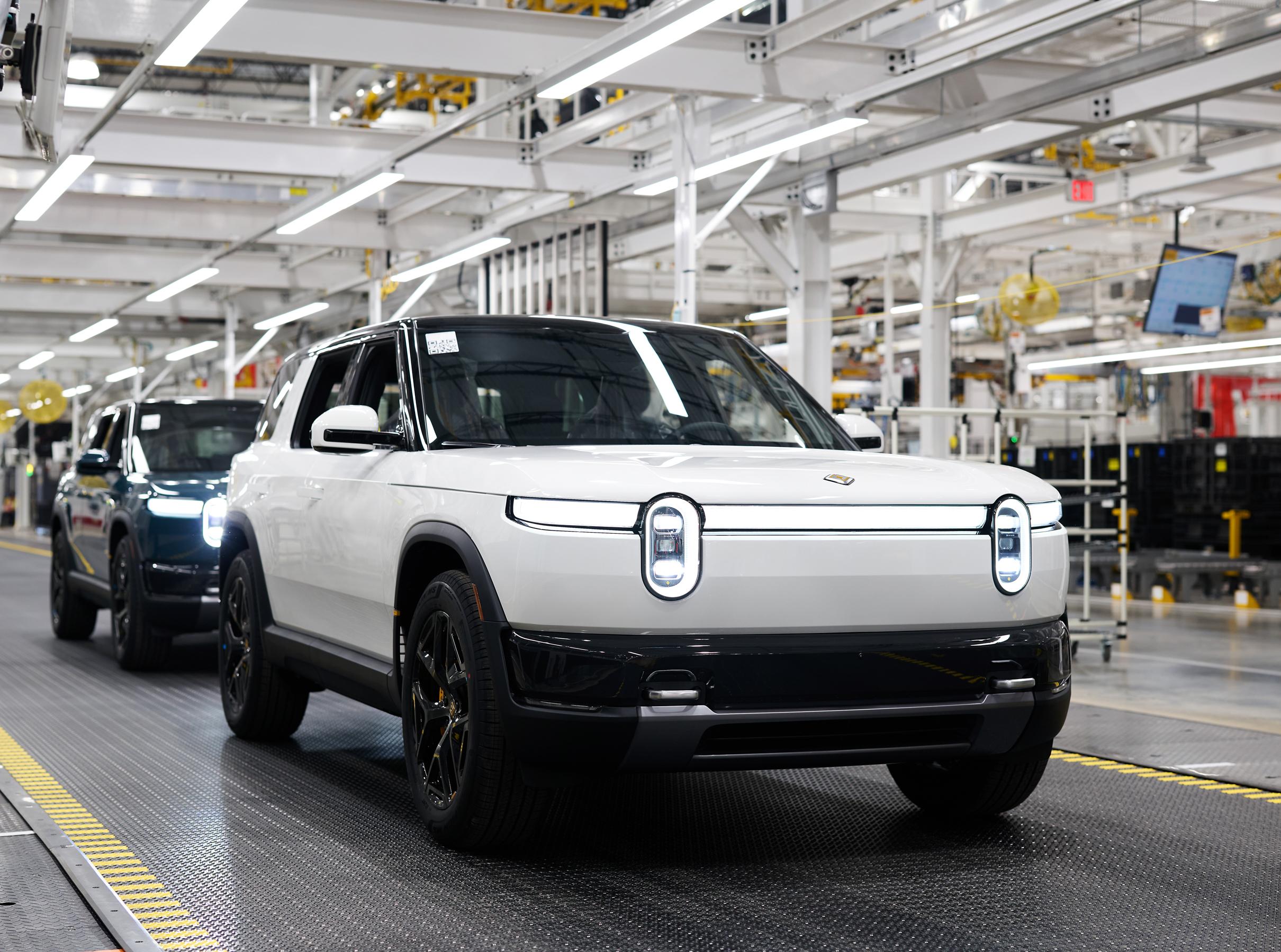 Two Rivian R2’s with headlights on are displayed, the first is white and the one behind is blue.