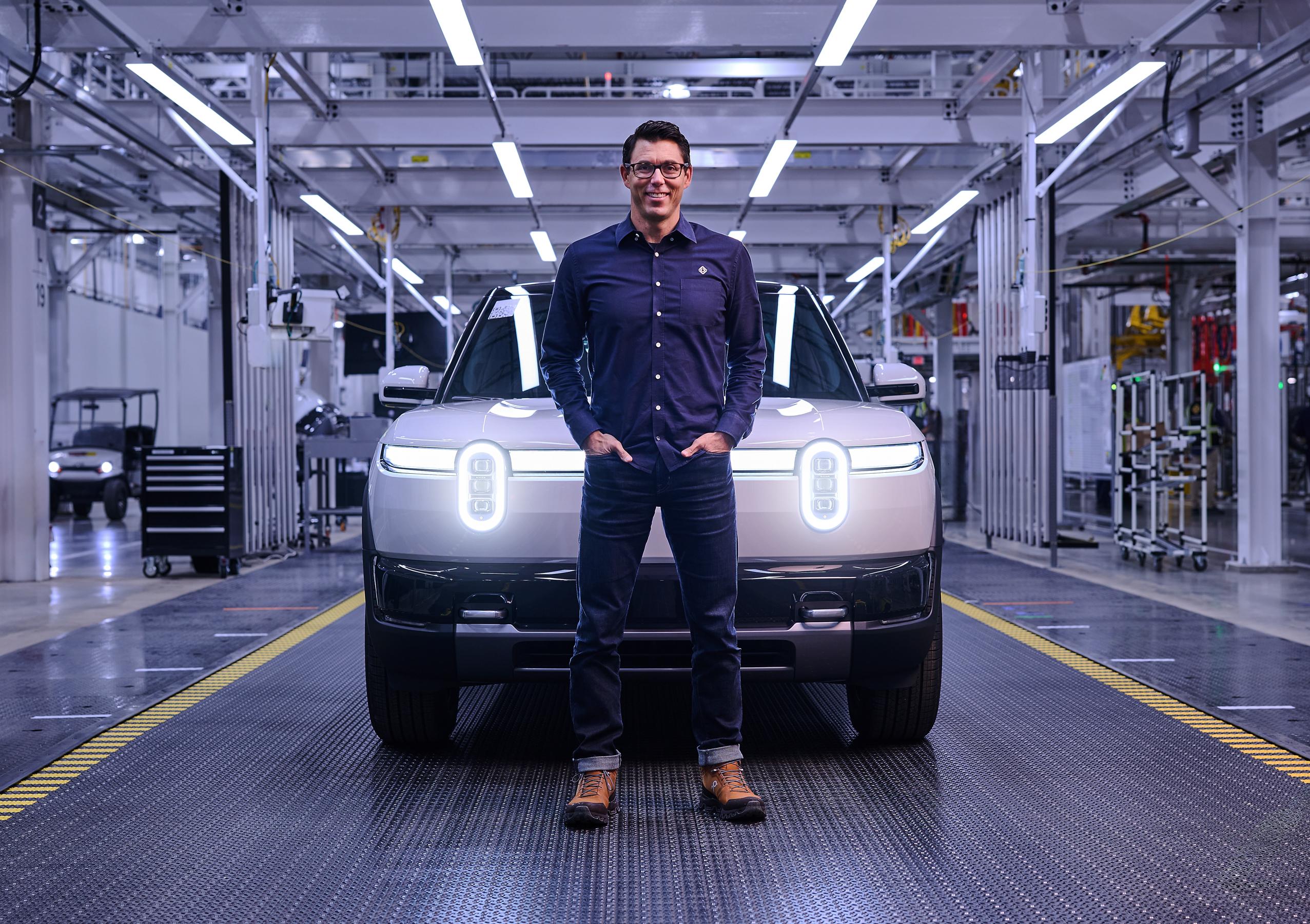 A smiling man wearing jeans, a blue button down shirt, boots and glasses stands in front of a white Rivian R2 with its headlights on. The image is taken in a factory and has a blue cast.