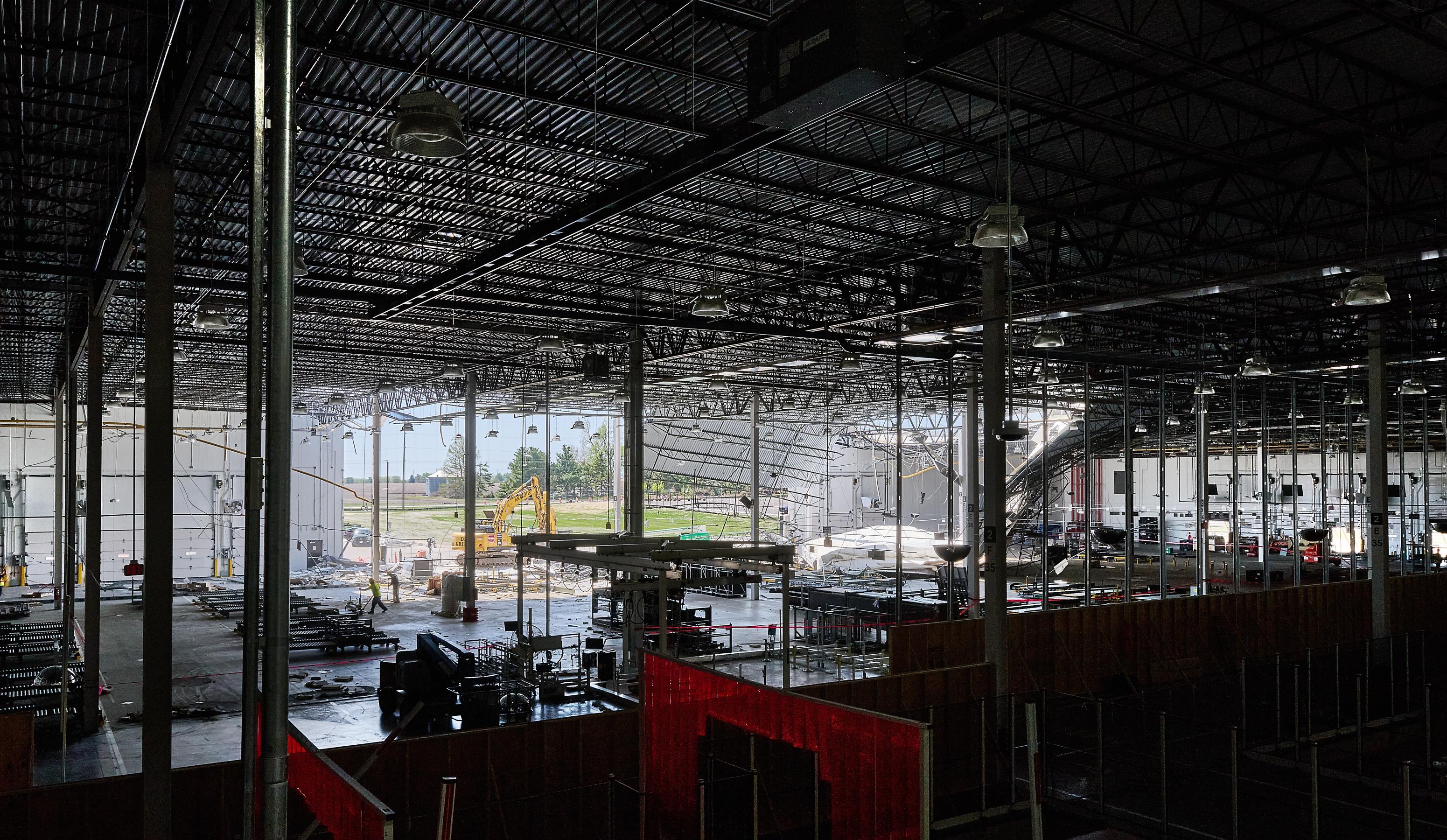 A wide view of a factory interior is mostly dark with a large open wall in the back where you can see to the bright daylight outside. A construction vehicle is in the center and debris can be seen hanging down.
